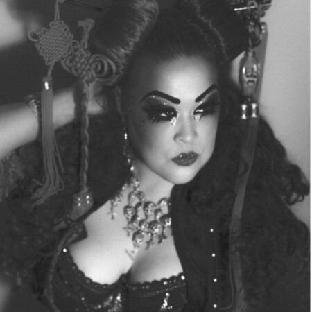 Lilly SnatchDragon, a person with dramatic make-up, bold eyebrows, and glittery eye shadow poses confidently. She wears elaborate hair accessories, large statement jewellery, and a sequinned outfit. The image is in black and white.