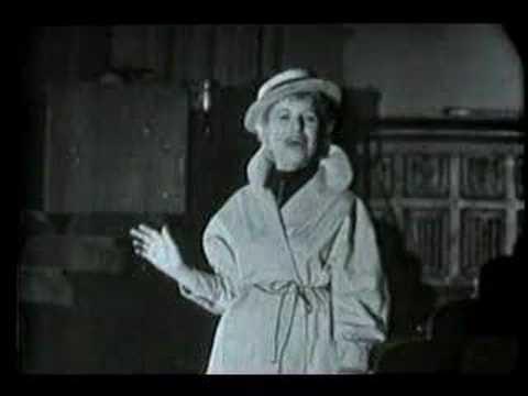 A woman in a coat and hat stands indoors, gesturing with one hand. The black-and-white image evokes a sense of history, suggesting it is from an older time period.