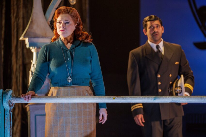 A woman with red hair and a blue jumper stands by a railing, looking sad. Behind her, a man in a dark naval officer’s uniform holds a white hat and looks concerned. The scene is dramatically lit.