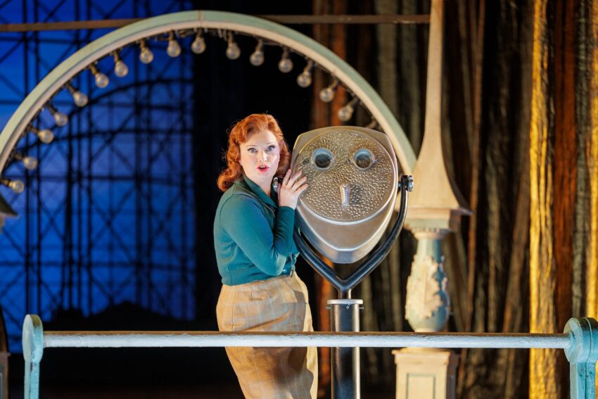 A woman with red hair and vintage clothing stands next to a large coin-operated binocular viewer, on a decorated stage with golden curtains and a lit arch in the background.