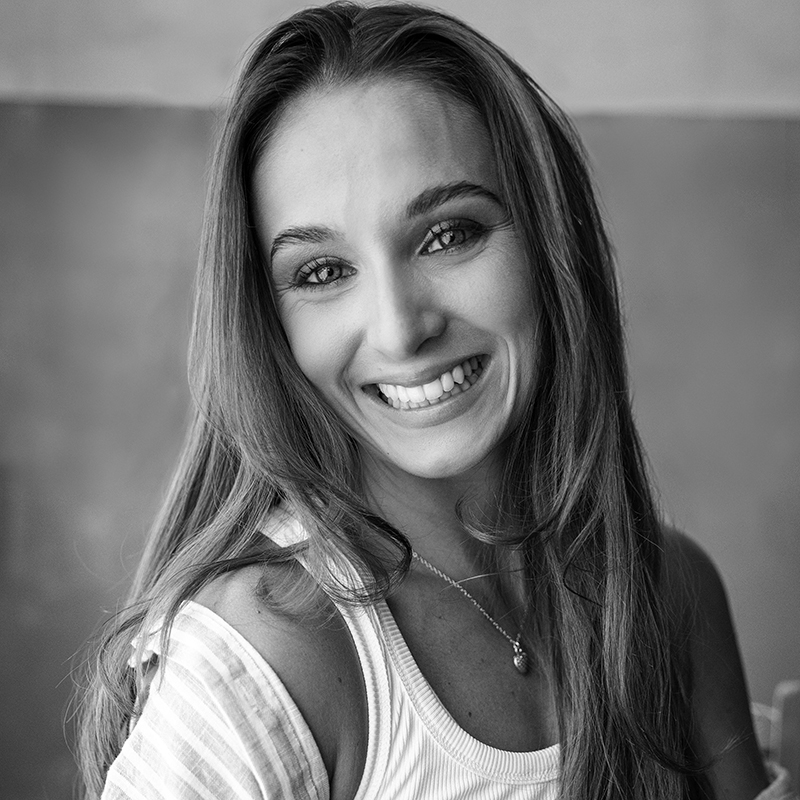 Megan Millie, with long hair, smiles brightly at the camera, wearing a sleeveless top and a necklace. The photo is in black and white, featuring a softly blurred background.