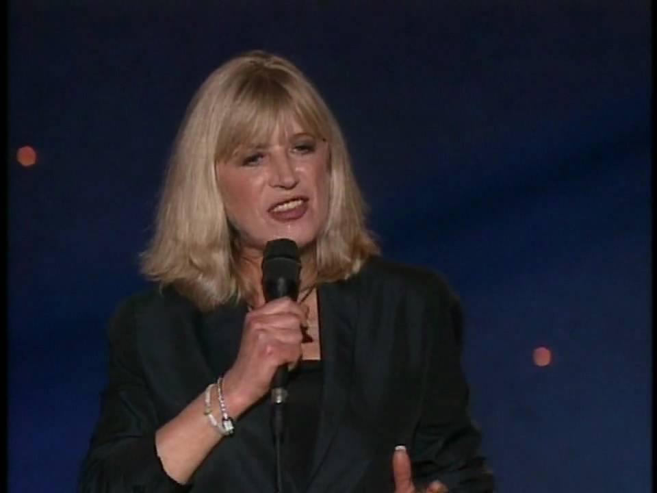 A woman with blonde hair wearing a dark outfit holds a microphone and speaks on stage against a dark blue background with small lights