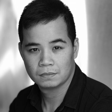 Black and white portrait of a young man with short dark hair, wearing a dark collared shirt, looking directly at the camera with a neutral expression. The background is softly blurred.