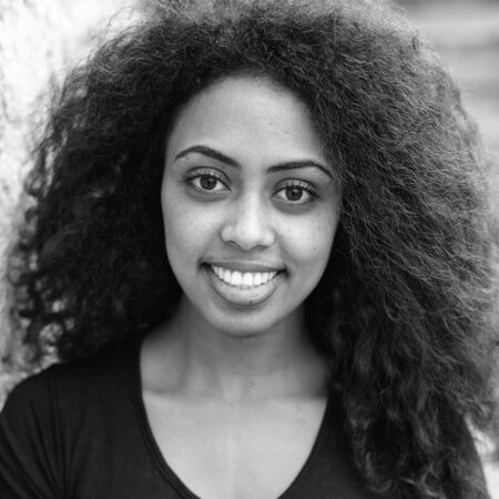 Senayt Aesfa Amare, a young woman with curly hair, smiles at the camera. She is wearing a dark shirt and is photographed in black and white, with a blurred background.