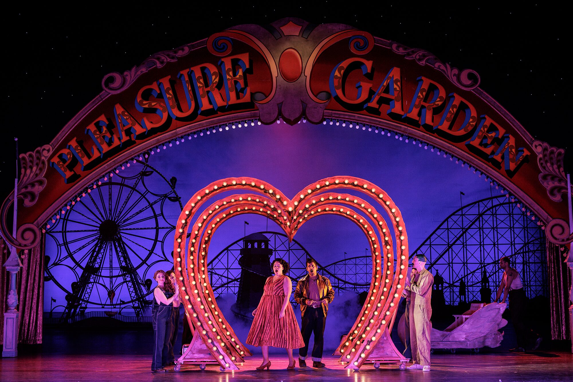 Actors perform on stage under a large Pleasure Garden sign, with a lit heart-shaped frame in the centre and amusement park rides, like a Ferris wheel and rollercoaster, depicted in the background.