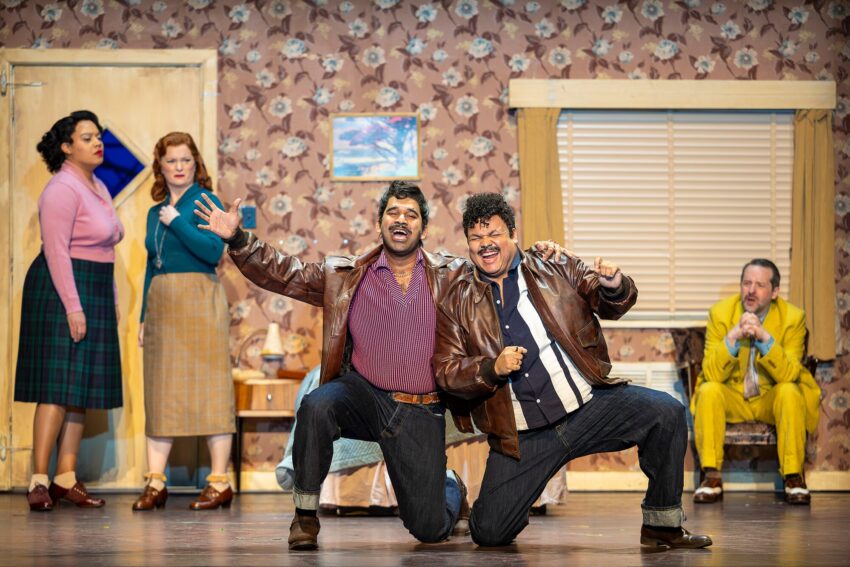 Five actors perform on stage in a retro-style room with floral wallpaper. Two men in brown jackets kneel and gesture dramatically at the front, while three others stand or sit in the background, reacting expressively.