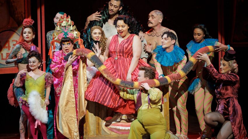 A group of actors in colourful, whimsical costumes perform on stage, surrounding a woman in a red dress who stands inside a large wooden mouth prop, creating a lively and theatrical scene.