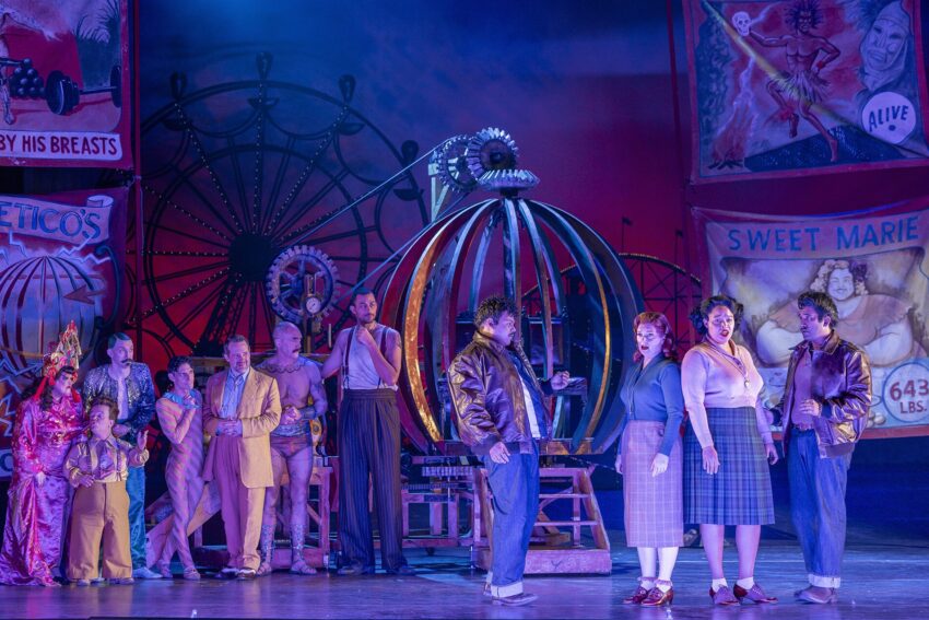 A group of performers in colourful costumes stand on stage in front of a large metal circus prop, with vintage sideshow banners and a Ferris wheel in the background. The lighting is dramatic and moody.