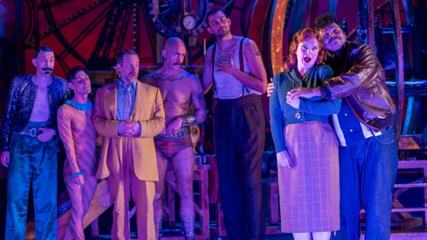 A group of seven people in colourful, theatrical costumes stand on stage under dramatic lighting. One man comforts a woman at the front, while five others watch with various expressions. The background features large gears and machinery.