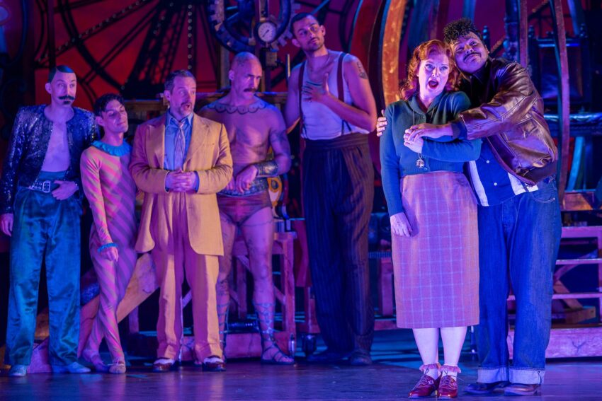 A group of seven people in colourful, theatrical costumes stand on stage under dramatic lighting. One man comforts a woman at the front, while five others watch with various expressions. The background features large gears and machinery.