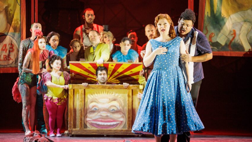 A group of performers in colourful costumes stand on stage, with a woman in a blue polka-dot dress singing in the foreground and a man holding her arm. Behind them is a carnival-themed set with painted banners and figures.