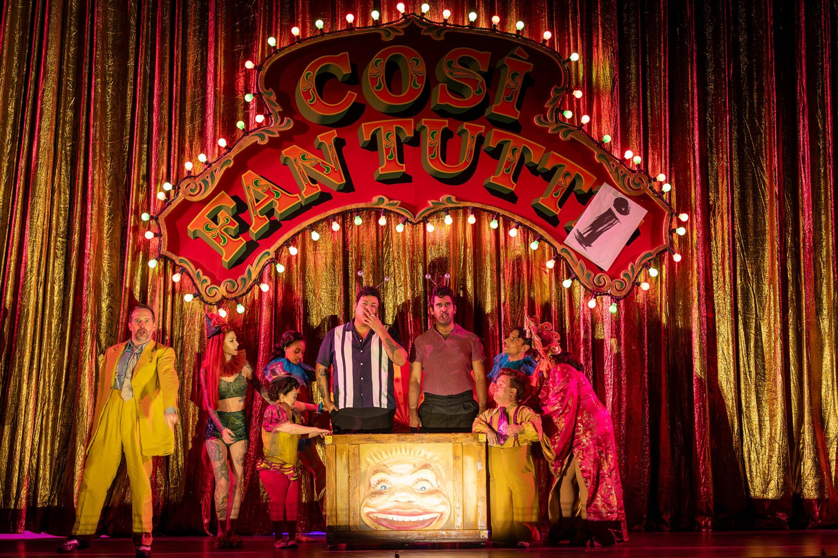 A colourful stage scene features performers in vibrant costumes standing in front of a glittering gold curtain and a large illuminated sign reading Così Fan Tutti. The actors pose animatedly around a wooden box with a clown face.