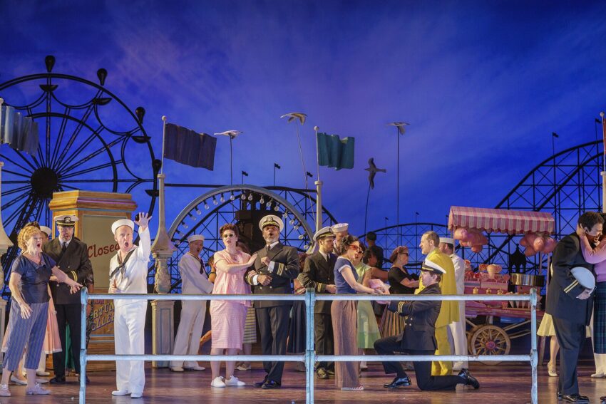 A lively stage scene set at a seaside amusement park, featuring performers in vintage costumes, including sailors and civilians, with rollercoasters and carnival stalls in the colourful background.