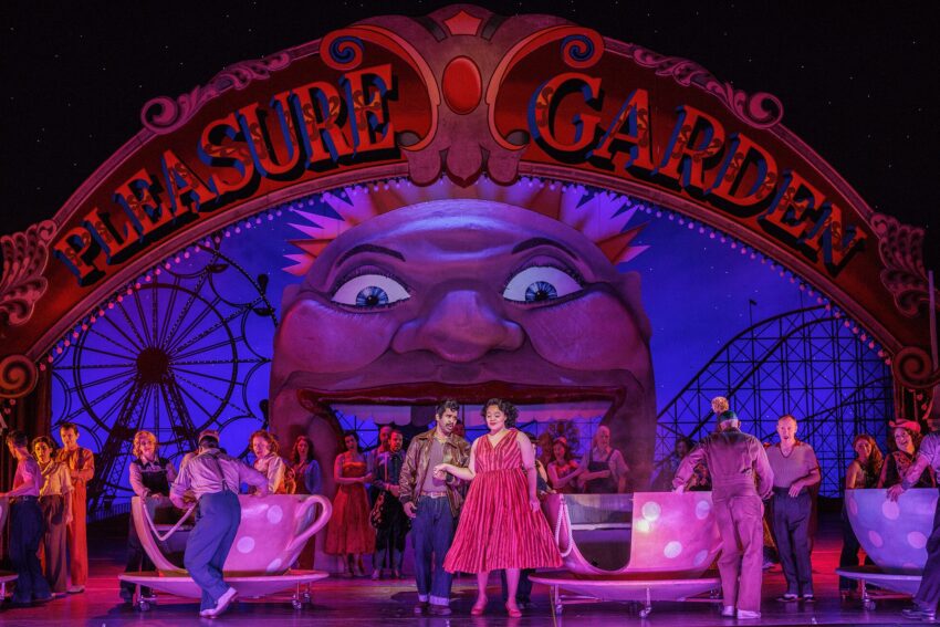 A vibrant stage scene of an opera set in a amusement park, featuring performers in colourful costumes, teacup ride props, and a large sign reading Pleasure Garden with a giant smiling face in the background.
