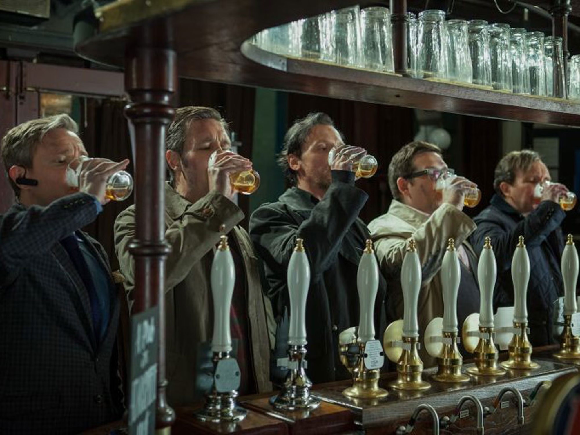 Five men stand in a row at a bar, each drinking from a pint glass of beer. Several white beer taps are in front of them and a shelf with glasses above their heads.