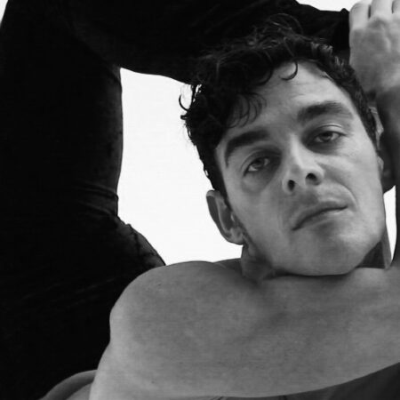 A man with curly hair, identified as Tommaso Di Vincenzo, holds an intense expression while performing an advanced flexibility pose, bringing his leg over his shoulder. The image is in black and white.