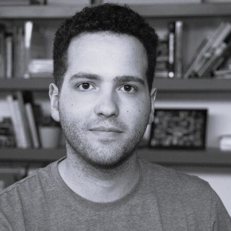 A young man with short curly hair and a light beard looks at the camera, sitting indoors in front of bookshelves filled with books and decorative items. This black and white portrait captures Ash J Woodward in a thoughtful moment.