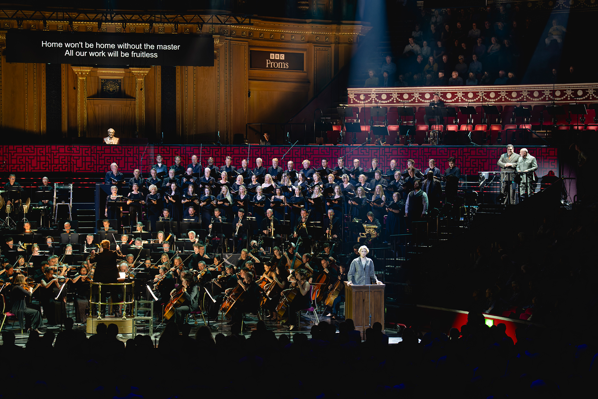 A conductor leads an orchestra and large choir onstage in a grand concert hall. A speaker stands at a lectern. Song lyrics are projected above: Home won’t be home without the master. All our work will be fruitless.