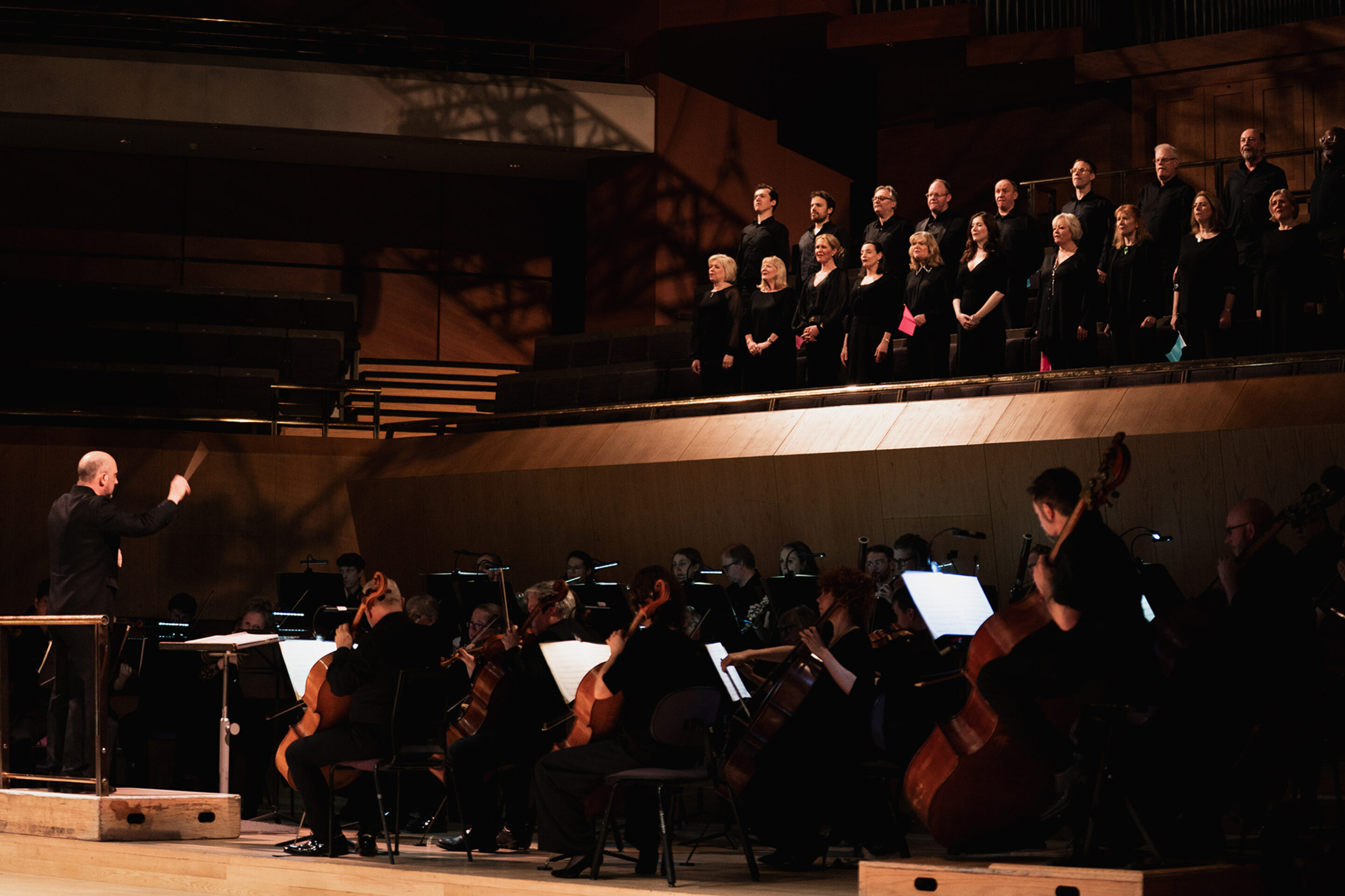 A conductor leads an orchestra playing string instruments while a choir stands on a raised platform behind them in a concert hall with warm lighting.