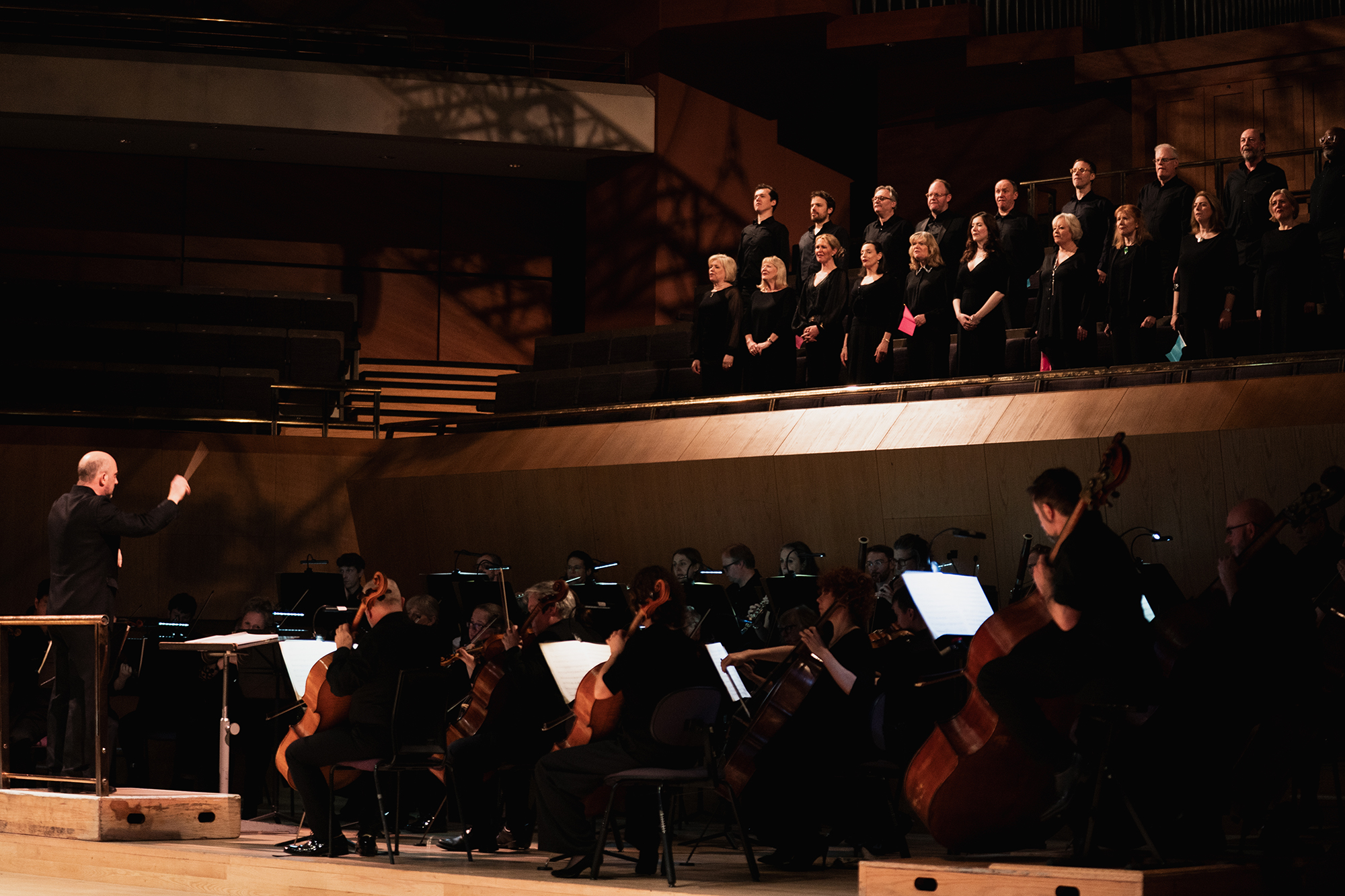 A conductor leads an orchestra playing string instruments while a choir stands on a raised platform behind them in a concert hall with warm lighting.