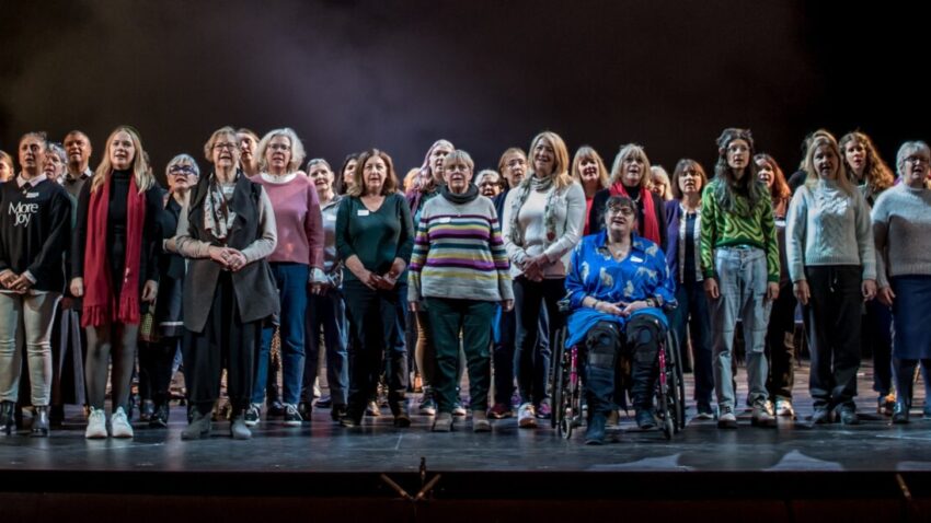 A large, diverse group of people stands on a theatre stage, facing the audience. Some are standing, one person is seated in a wheelchair, and tall set pieces resembling buildings promote Creative Health in the background.