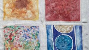 Four watercolour paintings on white fabric: top left is a brown and yellow abstract; top right is a red geometric star pattern; bottom left has colourful dots; bottom right shows blue and brown circles with vertical blue lines.