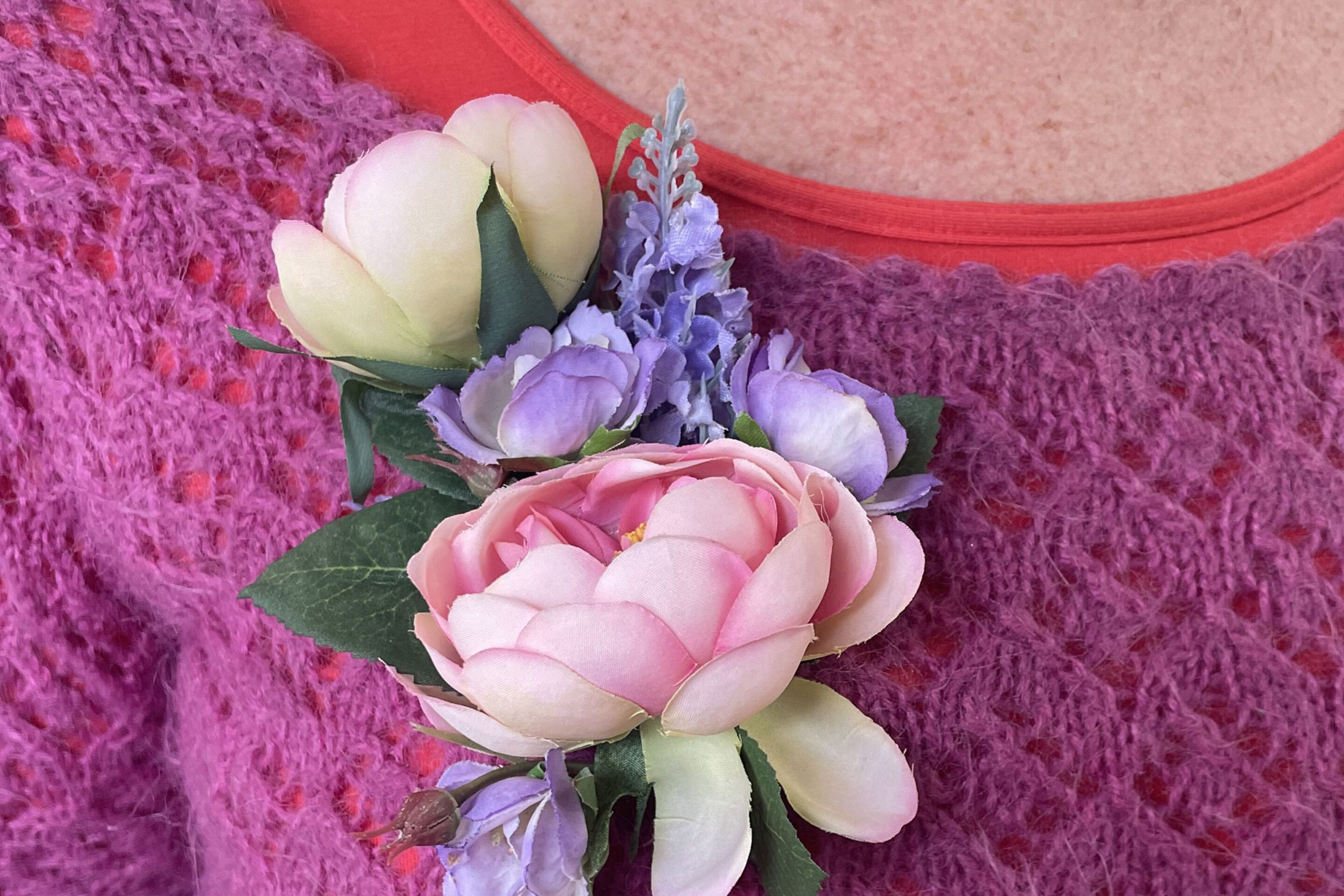 A close-up of a person wearing a knitted magenta jumper and red shirt, adorned with a floral corsage featuring pink and white flowers and green leaves pinned to the jumper.
