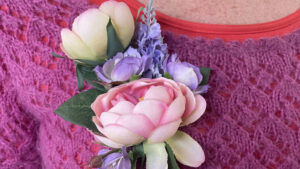 A close-up of a person wearing a knitted magenta jumper and red shirt, adorned with a floral corsage featuring pink and white flowers and green leaves pinned to the jumper.