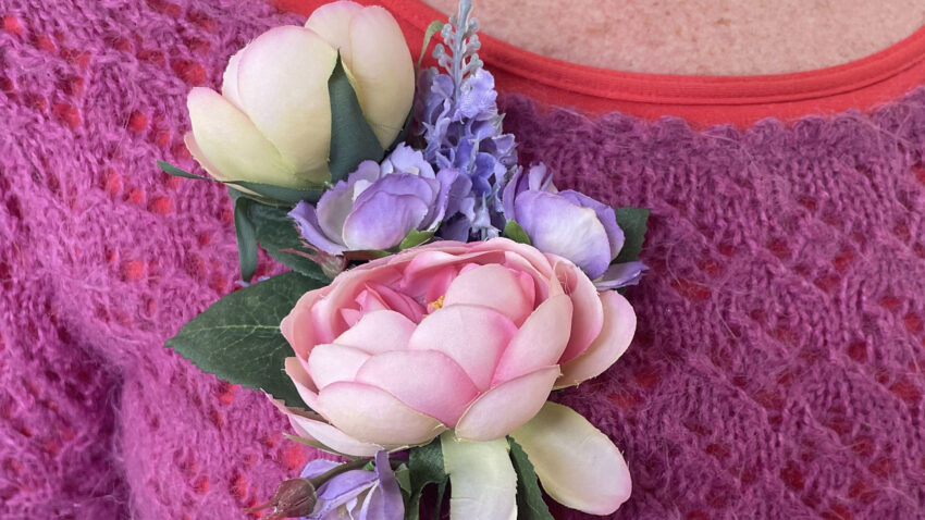 A close-up of a person wearing a knitted magenta jumper and red shirt, adorned with a floral corsage featuring pink and white flowers and green leaves pinned to the jumper.