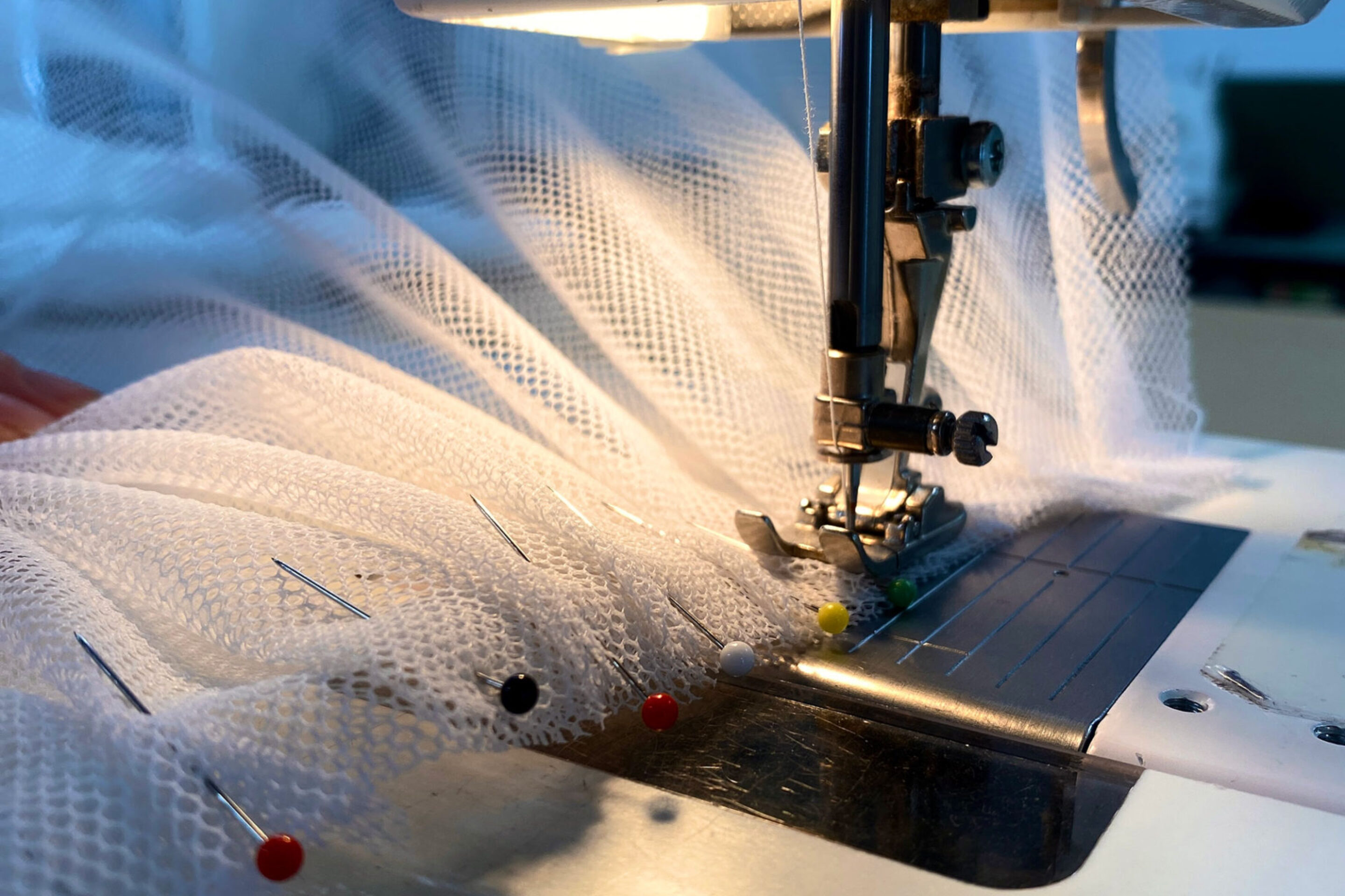 Close-up of a sewing machine stitching white mesh fabric, with several colourful pins securing the material in place under the needle.