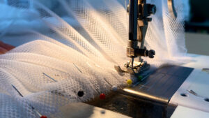Close-up of a sewing machine stitching white mesh fabric, with several colourful pins securing the material in place under the needle.
