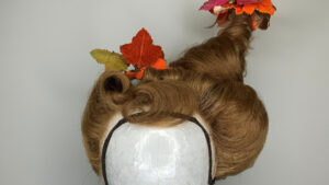 A wig styled in an elaborate updo with a tall, twisted peak, decorated with artificial autumn leaves in red, orange, and yellow. The wig is displayed on a white mannequin head against a plain background.