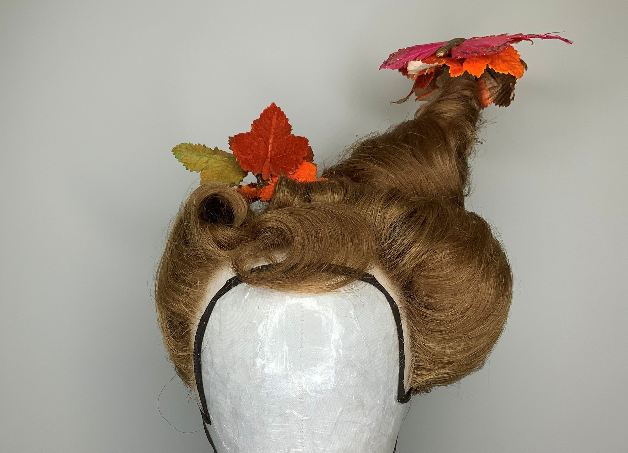 A wig styled in an elaborate updo with a tall, twisted peak, decorated with artificial autumn leaves in red, orange, and yellow. The wig is displayed on a white mannequin head against a plain background.