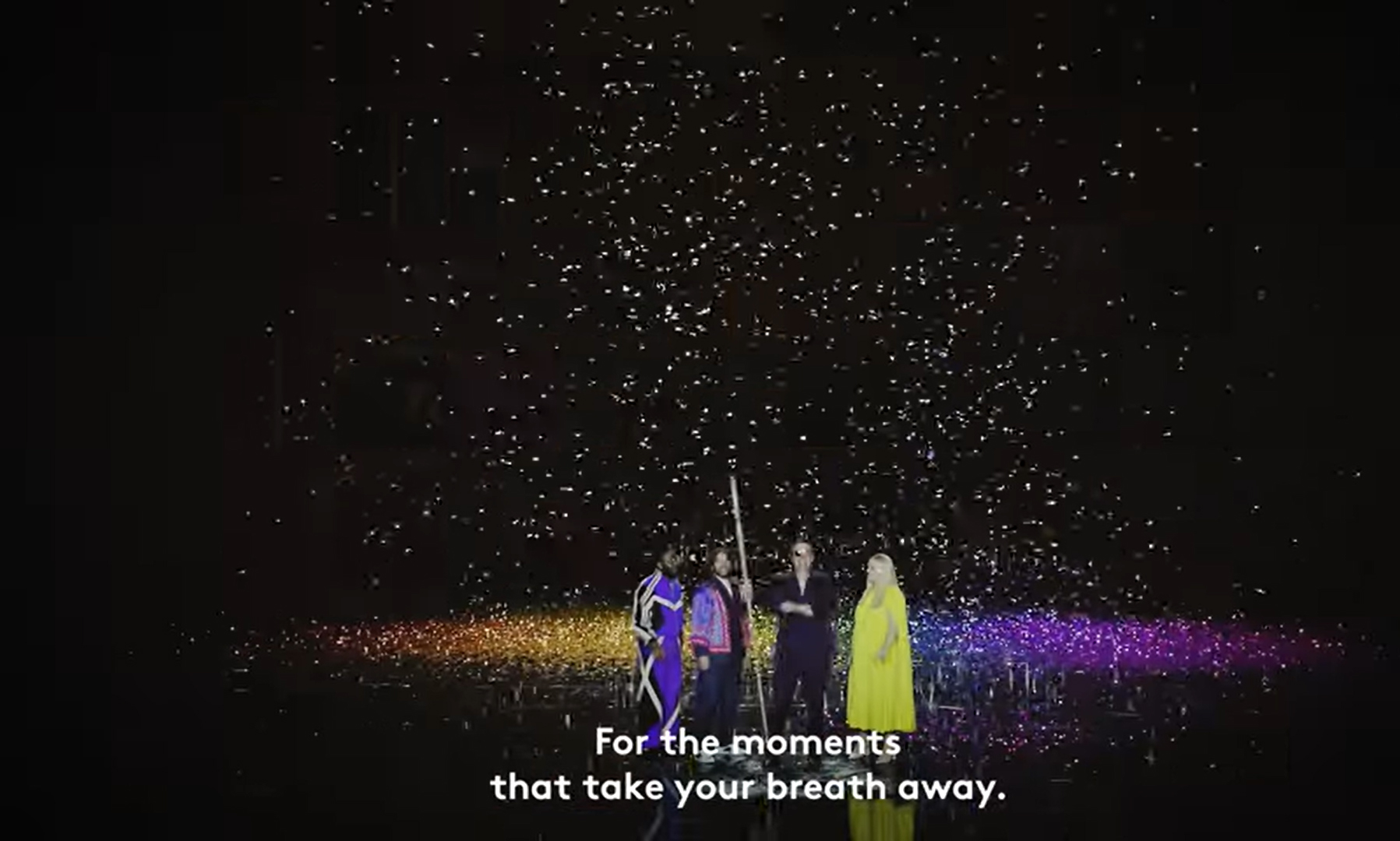 Three people stand under a shower of sparkling lights on a dark stage, with rainbow colours at their feet. White text at the bottom reads, For the moments that take your breath away.