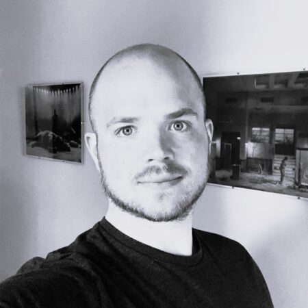 Jack Knowles, a man with a shaved head and trimmed beard, smiles slightly in a black-and-white selfie. He wears a dark T-shirt indoors, with two framed photos hanging on the plain wall behind him.