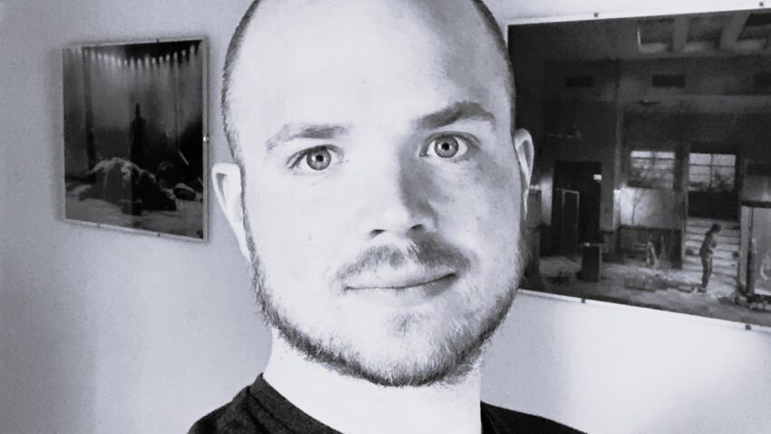 Jack Knowles, a man with a shaved head and trimmed beard, smiles slightly in a black-and-white selfie. He wears a dark T-shirt indoors, with two framed photos hanging on the plain wall behind him.