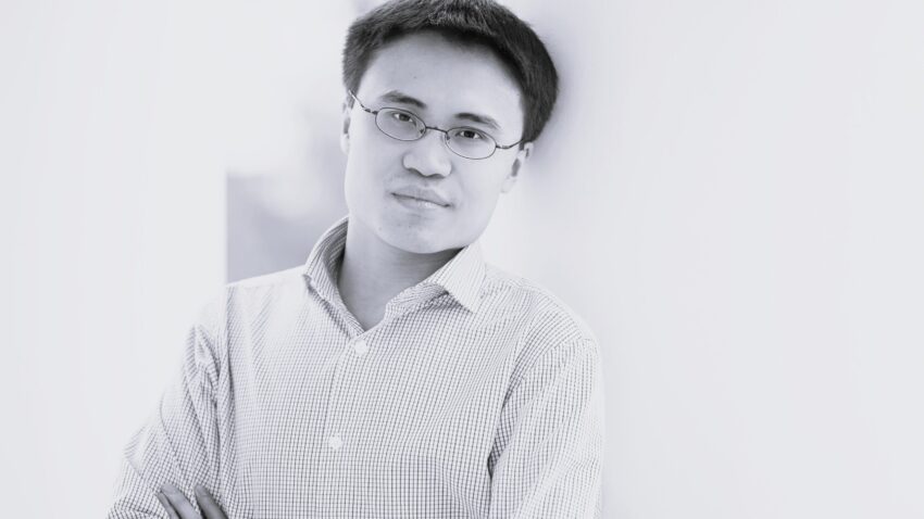 A person with short hair and glasses leans against a white wall, arms folded, wearing a light-coloured checked shirt, and looking at the camera with a neutral expression. The background is softly blurred.