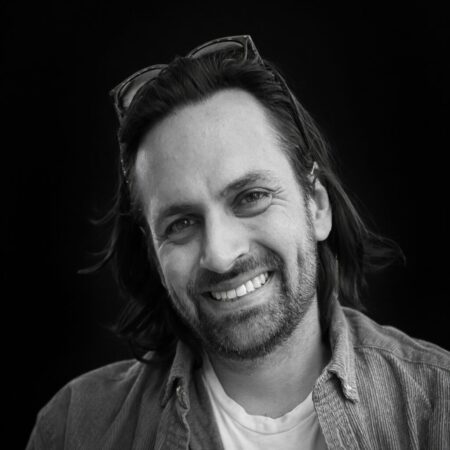 Black and white portrait of Joseph Reiser, a smiling man with medium-length hair, a beard, and moustache. He wears a casual shirt over a T-shirt, with sunglasses resting on his head, set against a dark background.