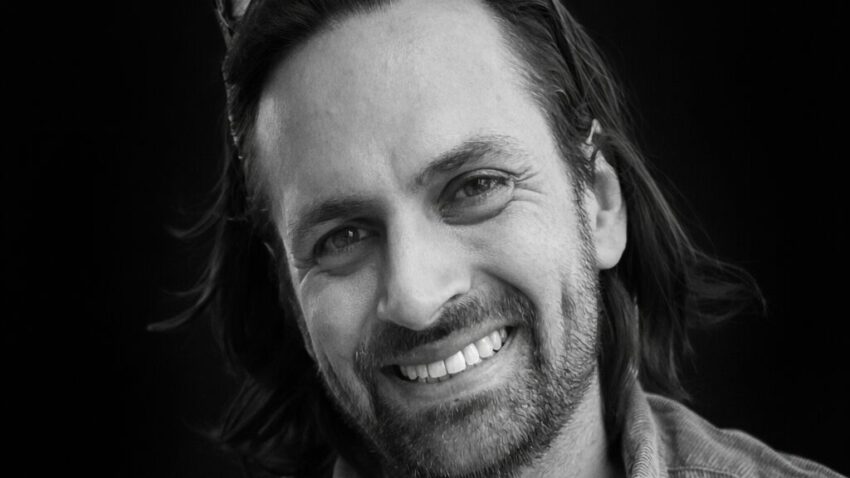 Black and white portrait of Joseph Reiser, a smiling man with medium-length hair, a beard, and moustache. He wears a casual shirt over a T-shirt, with sunglasses resting on his head, set against a dark background.