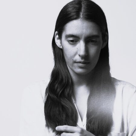 A woman with long, dark hair and a serious expression looks down, wearing a light-coloured shirt against a plain, light background. The black and white image captures Mariam Wallentin's contemplative mood.