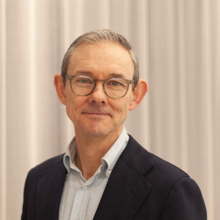 A middle-aged man with short grey hair and glasses, Paul Reeve MBE is wearing a navy blazer over a light blue shirt, standing in front of pale-coloured curtains and smiling slightly at the camera.