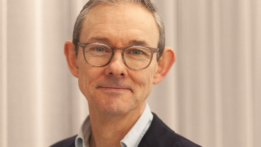 A middle-aged man with short grey hair and glasses, Paul Reeve MBE is wearing a navy blazer over a light blue shirt, standing in front of pale-coloured curtains and smiling slightly at the camera.