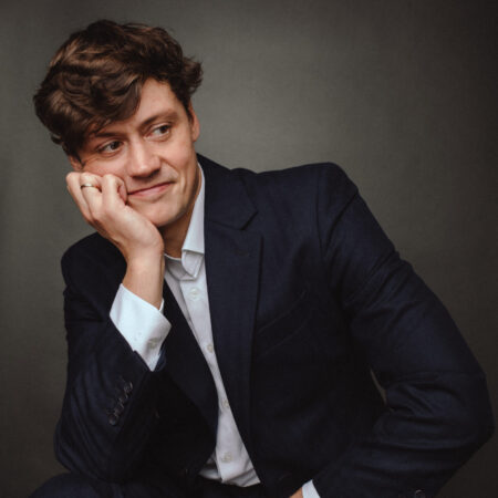 A man in a dark suit and white shirt sits with his chin resting on his hand, smiling and looking to the side against a plain dark background.