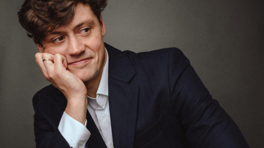 A man in a dark suit and white shirt sits with his chin resting on his hand, smiling and looking to the side against a plain dark background.
