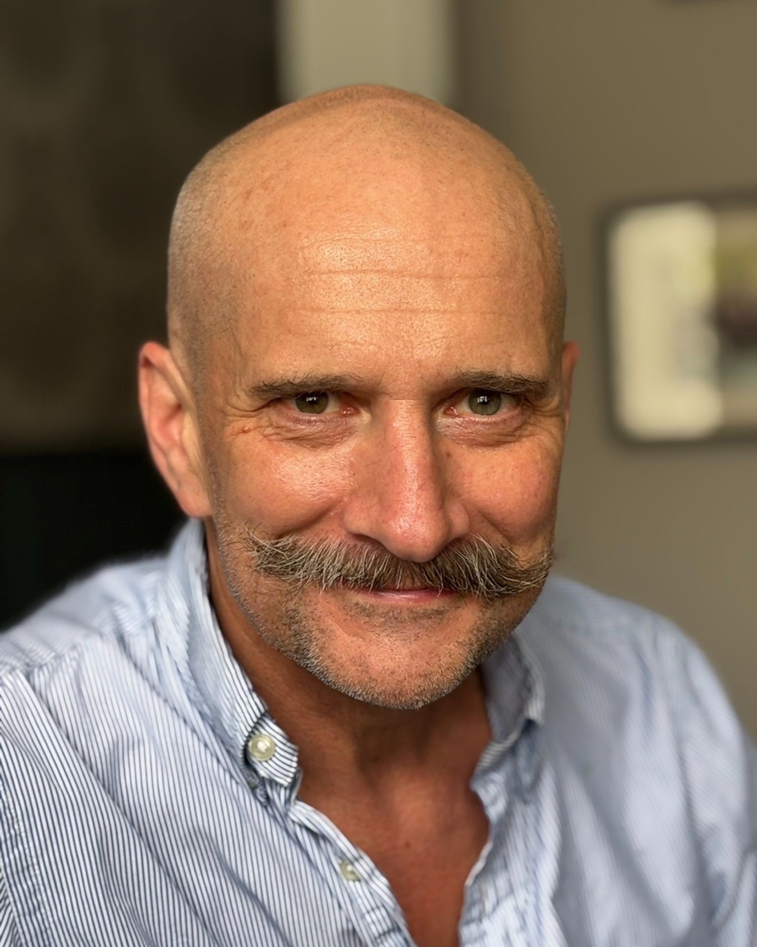Andy, a middle-aged man with a bald head and handlebar moustache, smiles at the camera in a light blue, button-up shirt. The softly blurred background with neutral tones gives the photo a warm, friendly vibe—perfect for a chat.
