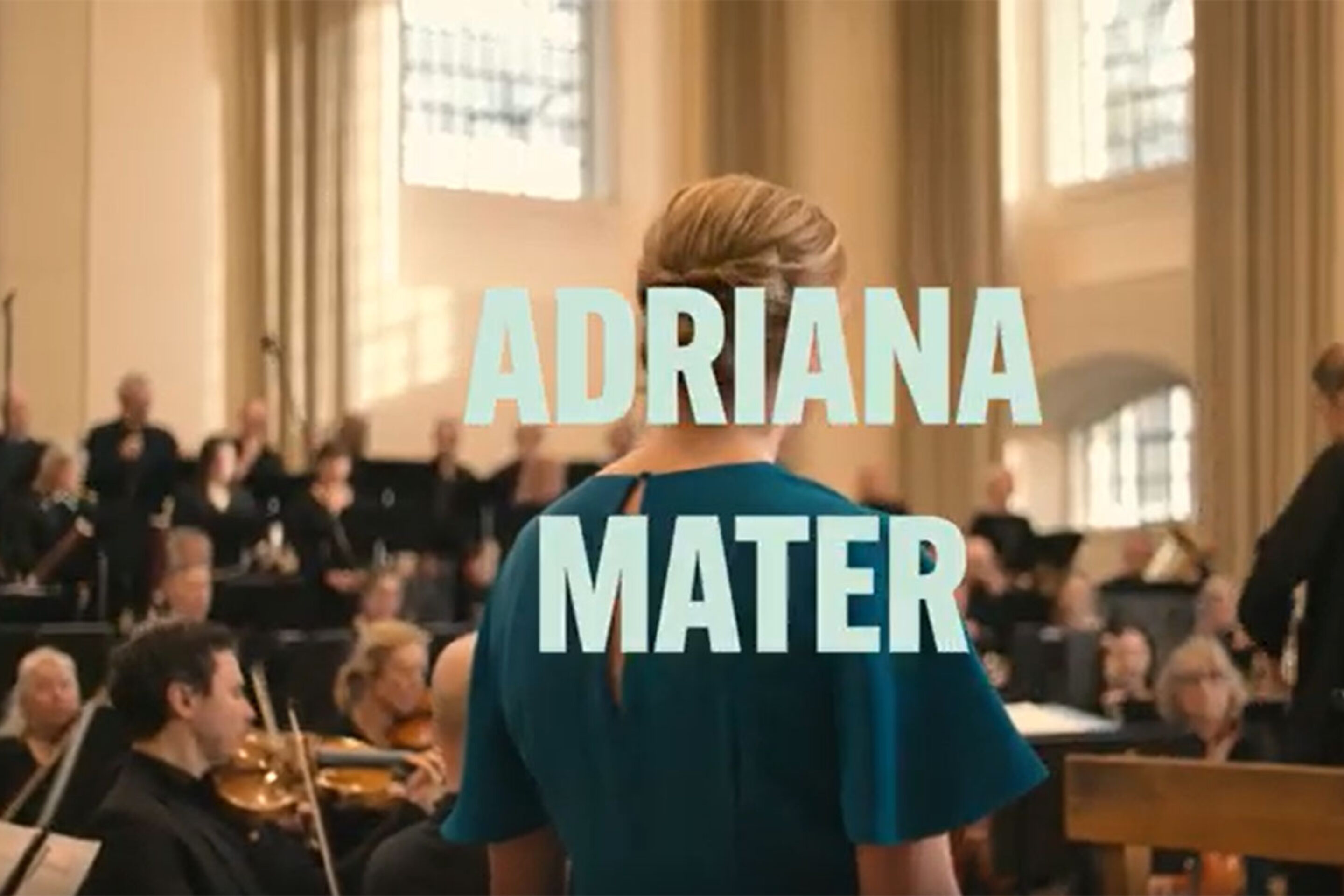 A woman in blue stands before an orchestra and conductor in a bright concert hall. Large text over her reads ADRIANA MATER, evoking Kaija Saariaho’s powerful opera.