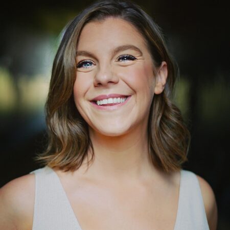 Alison Langer, with shoulder-length light brown hair and blue eyes, smiles brightly in a light-coloured sleeveless top. The background is blurred and dark, drawing focus to her face.