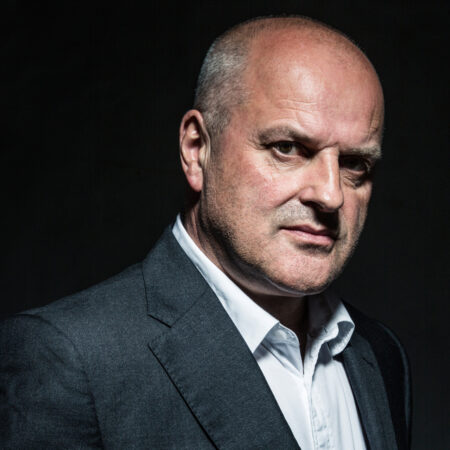 Christopher Purves, a middle-aged man with a bald head, wears a dark grey suit jacket and white shirt, looking seriously into the camera against a dark background.