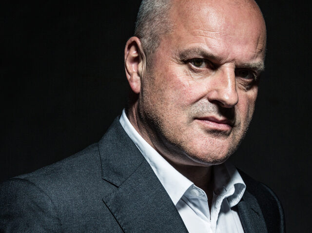Christopher Purves, a middle-aged man with a bald head, wears a dark grey suit jacket and white shirt, looking seriously into the camera against a dark background.