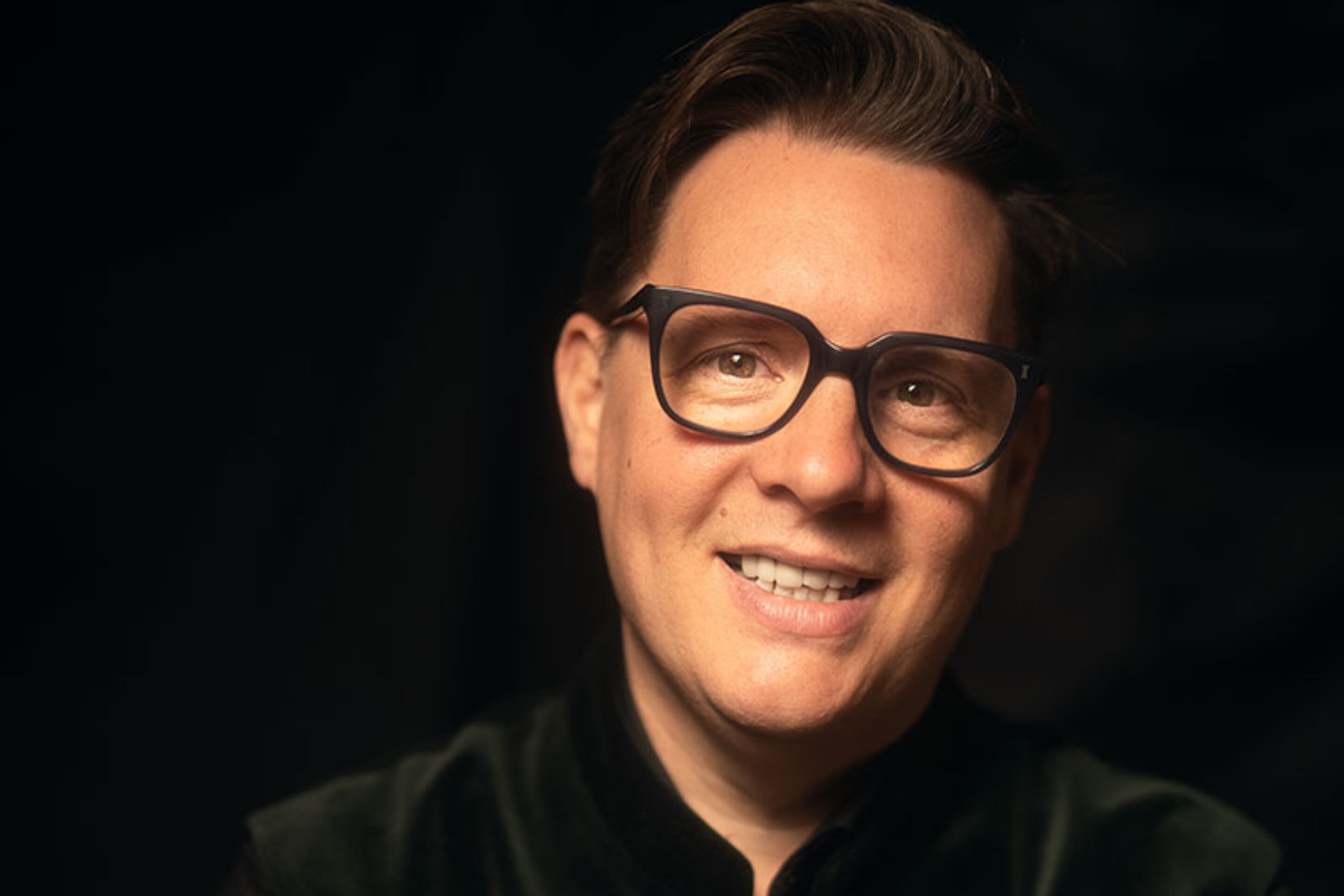 A smiling person with short brown hair, wearing glasses and a dark, buttoned-up velvet jacket, sits against a dark, softly lit background.